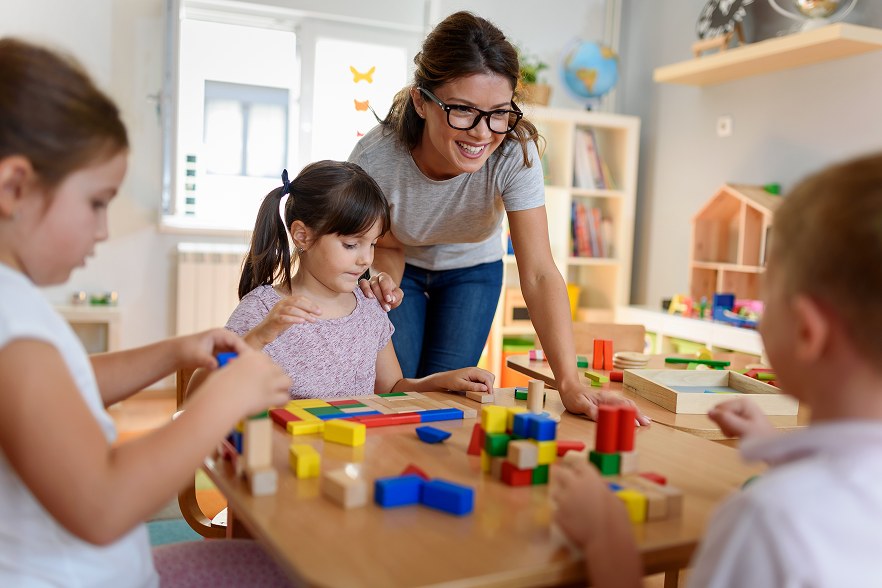 Éducatrice souriante accompagnant de jeunes enfants qui jouent avec des cubes en bois colorés autour d'une table