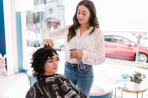 Coiffeuse vaporisant les cheveux bouclés d'une cliente assise sous une cape, près de la vitrine d'un salon de coiffure
