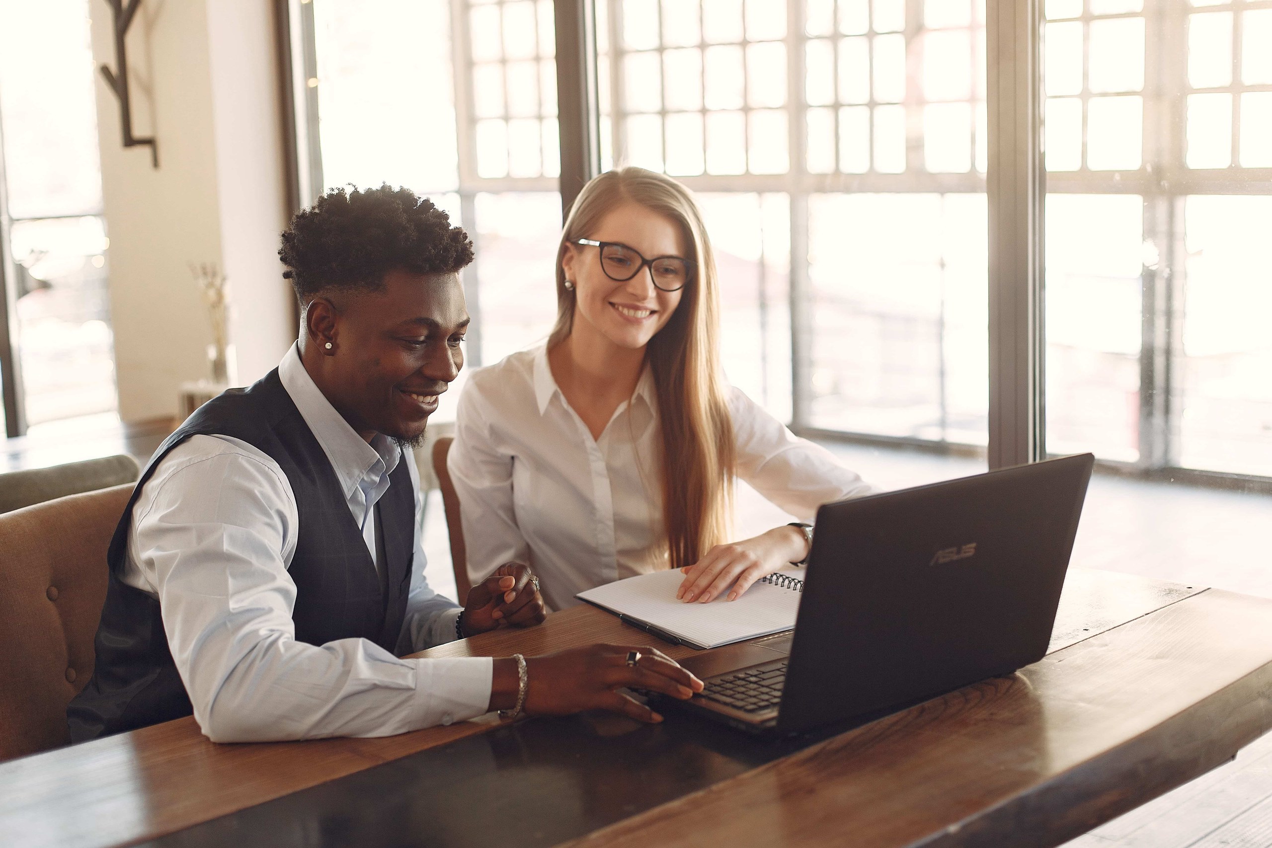 Deux entrepreneurs souriants assis côte à côte consultant un ordinateur portable et un carnet de notes sur une table en bois