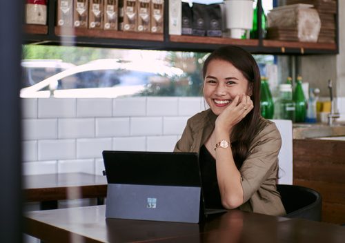 Commerçante souriante assise derrière une tablette à une table de son café, avec des étagères de produits au fond