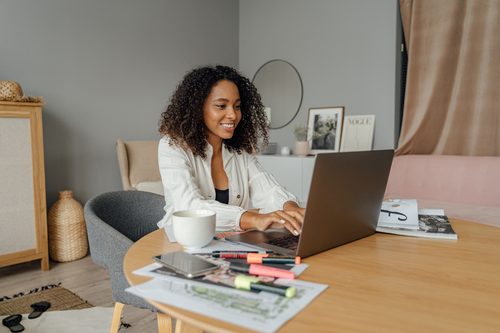 Femme souriante travaillant sur un ordinateur portable entourée de documents, surligneurs et d'une tasse de café