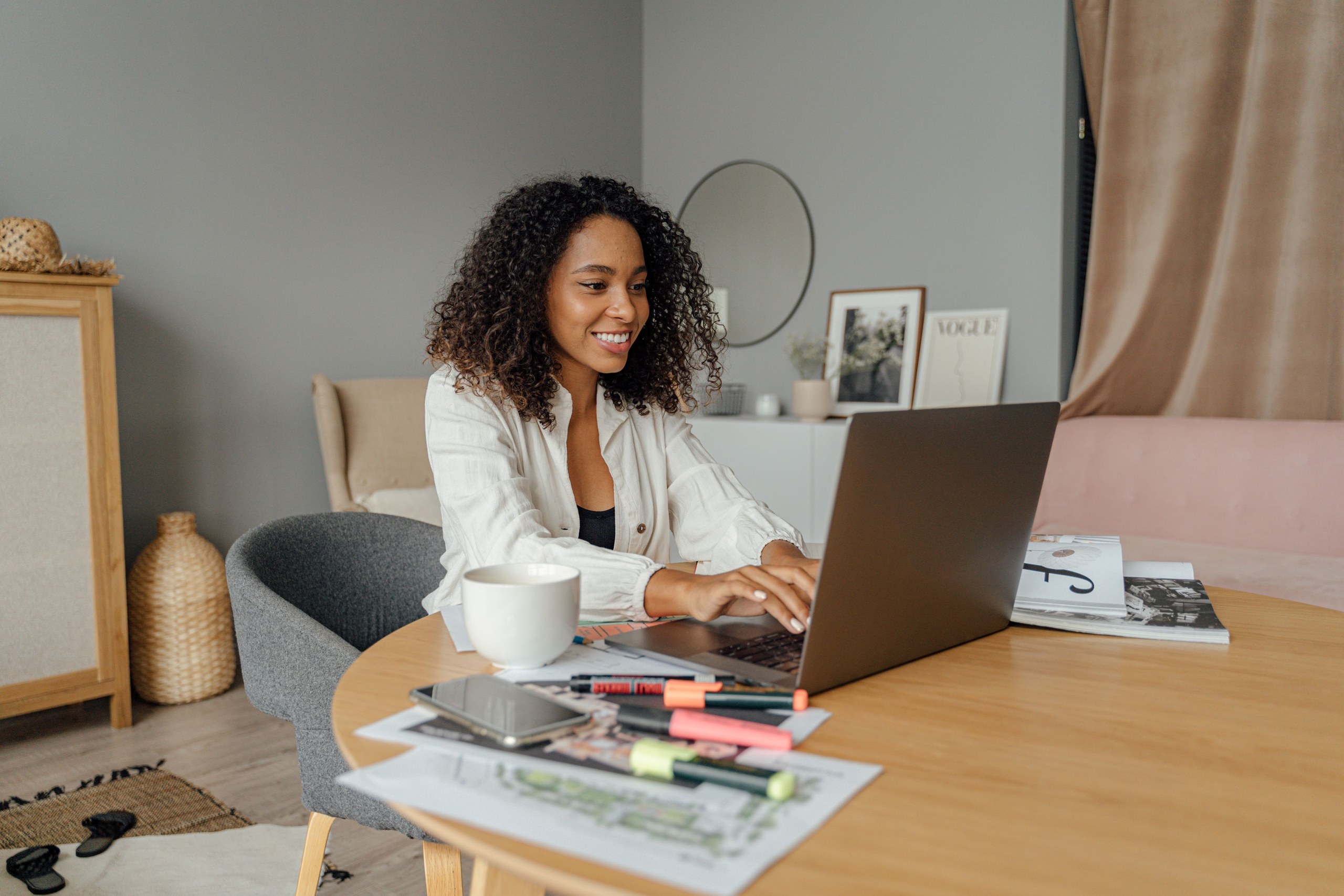 Femme souriante travaillant sur un ordinateur portable entourée de documents, surligneurs et d'une tasse de café
