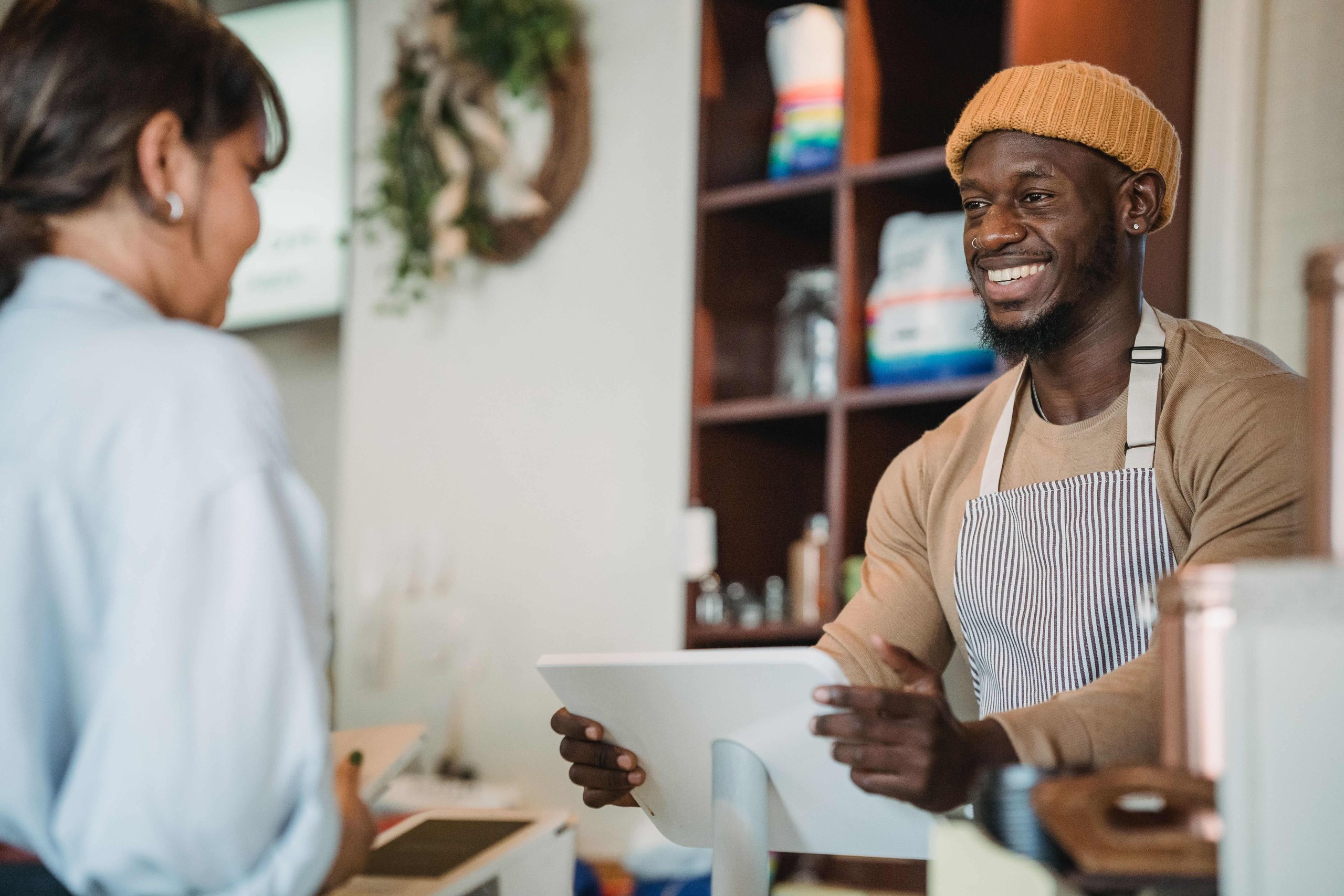 Commerçant souriant en tablier rayé encaissant une cliente sur une caisse tablette au comptoir de sa boutique