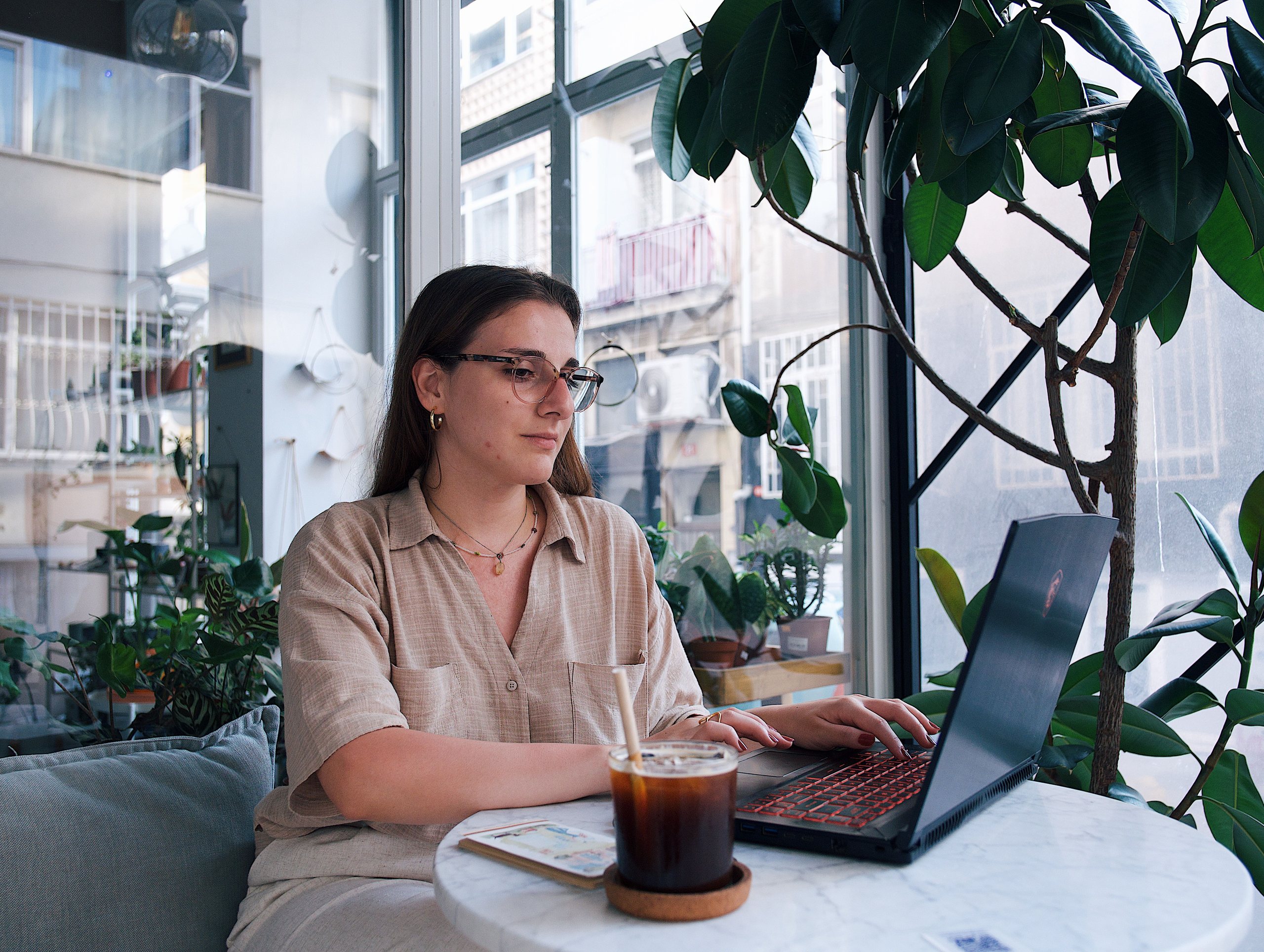 Jeune femme à lunettes travaillant sur un ordinateur portable à une table de café lumineux entourée de plantes vertes